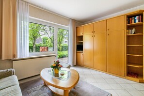 Ein Wohnzimmer mit einer Couch, einem Glastisch mit einer Vase, einem Bücherregal aus Holz und einem großen Fenster mit Vorhängen.