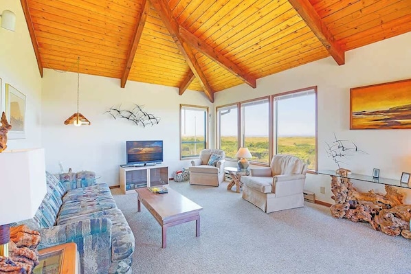 A spacious living area with a high wooden ceiling, large windows, a television, and a sofa.