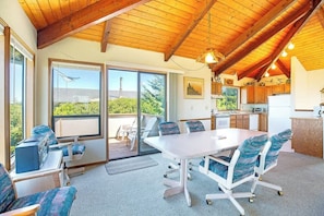 A spacious room with a large table and chairs, a kitchen area, and large windows offering a view of greenery.