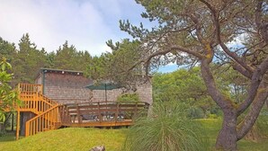 Una cabaña rústica con terraza y escalera de madera, rodeada de abundante vegetación y árboles.