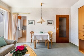 Ein gemütlicher Wohnbereich mit Essplatz, einer Couch, einem Couchtisch mit Blumen, einer Holztür und einer Küche im Hintergrund.