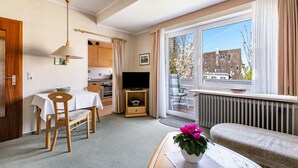 Ein Zimmer mit Esstisch, Stühlen, Fernseher, Sofa, Fenster mit Blick auf Gebäude und Heizkörper.