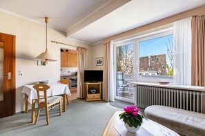Une pièce comprenant une table à manger, des chaises, un téléviseur, un canapé, une fenêtre donnant sur des immeubles et un radiateur.