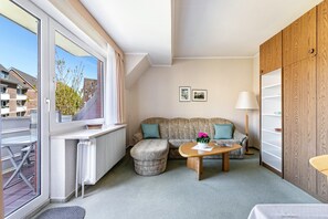 Un salon moderne comprenant un canapé, un pouf et une table basse, un radiateur, une fenêtre donnant sur des immeubles et une armoire en bois.