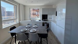 Une cuisine moderne avec des meubles blancs, une table à manger dressée pour quatre personnes et une vue sur le paysage urbain, visible à travers de grandes fenêtres.