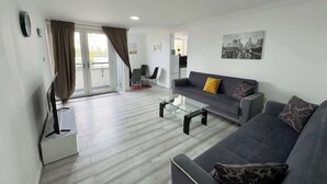A modern living room with a grey sofa, a glass coffee table, and a cityscape painting on the wall. - Riverview Stunning Apartment 3Bedroom 2Bathroom (Greater London)