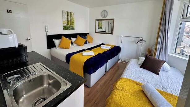 Camera d'albergo con angolo cottura, due letti con lenzuola gialle e blu, una sveglia e una finestra con tende.