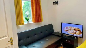 Una habitación con un sofá negro, un televisor mostrando una receta de galletas y una ventana con cortinas.