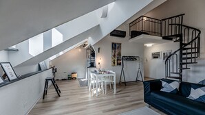 A modern loft apartment with a dining area, a dark blue sofa, a staircase, and a view of the city. - [Historic Palace Duplex] Cruise 2 min • A/C • WiFi (Savona)
