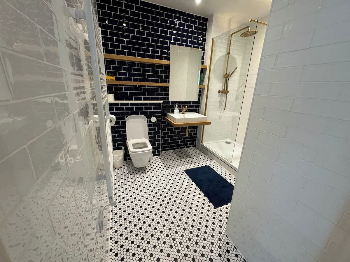 Une salle de bain avec un carrelage noir et blanc au sol, des toilettes blanches, une cabine de douche en verre et un lavabo blanc avec un miroir au-dessus.