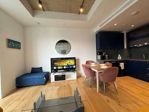 A modern living space with a sofa, TV, dining table, chairs, and a kitchen area. - London Cosy flat Balcony, RiverView, O2, Excel (Greater London)