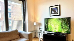 Ein modernes Wohnzimmer mit großem Flachbildfernseher, einer Couch, einem Glastisch und einem gerahmten Bild an der Wand.