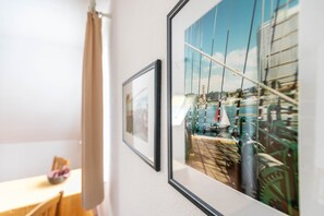 Framed artwork on a wall, a wooden table with a chair, and a window with a view of a dock and boats. - Apartment "No. 10" with Wi-Fi (Büsum)