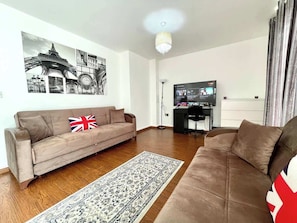 Ein Wohnzimmer mit einer Couch, einem Teppich, einem Fernseher und einem Schreibtisch mit Computer.