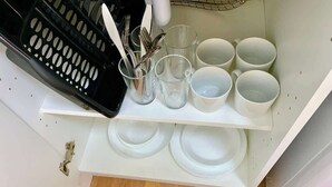 Una cocina con encimera blanca, una cesta de frutas negra y varias tazas y platos blancos.