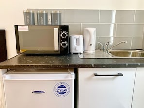 A kitchenette with a microwave, coffee maker, and tea maker on the counter, a sink, and a white refrigerator with a 2-year guarantee sticker. - Studio flat en-suite, Kitchenette, Parking, NP3 (Newbury Park)