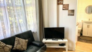 A living room with a black sofa, a TV on a white stand, a glass table, and a bathroom visible through an open door. - En-suite Studio Room with Kitchenette, Parking (Greater London)