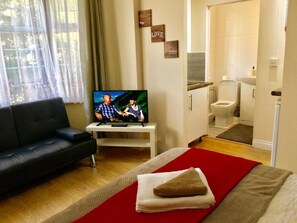 A room with a TV on a white stand, a black sofa, a bed with a red and beige blanket, and a bathroom visible through an open door. - En-suite Studio Room with Kitchenette, Parking (Greater London)