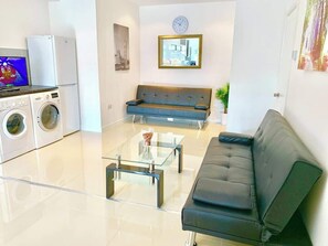 A modern living room with a sofa, a glass coffee table, a washing machine, and a clock on the wall. - 2 Bedroom Ground Floor House, Garden and Parking (Newbury Park)