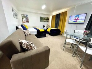 Ein Hotelzimmer mit einer Couch, einem Bett mit gelb-blauer Bettwäsche, einem Glastisch mit Stühlen und einem Fernseher an der Wand.