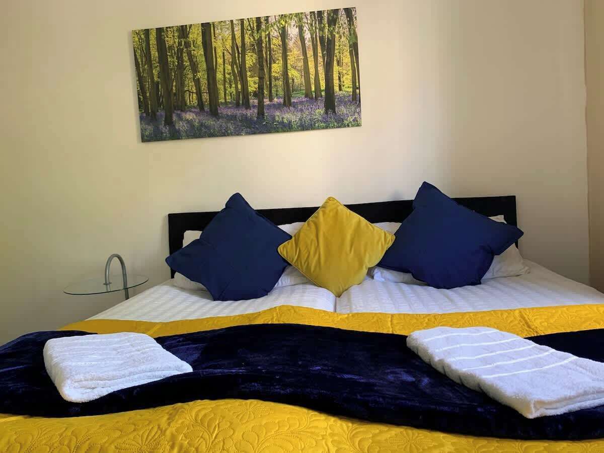 A bed with a yellow and blue bedspread, two pillows, a dark blue headboard, and a painting of a forest on the wall.