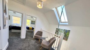 Ein Zimmer mit hoher und schräger Decke, einer Treppe, zwei Sesseln, einem kleinen Tisch, einer Chandelier und einem Gemälde.