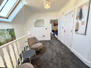 Un couloir avec un sol recouvert de moquette, une chaise, une petite table, un luminaire au plafond et une porte.