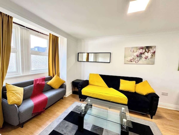 A modern living room with two sofas, one grey and one yellow, a glass coffee table, and a painting on the wall.