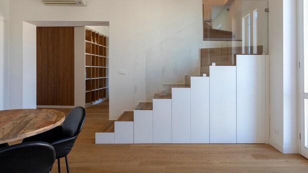 Un intérieur moderne avec une table à manger en bois, des chaises noires, un escalier avec des rampes en verre et un mur blanc agrémenté d’un panneau en bois.