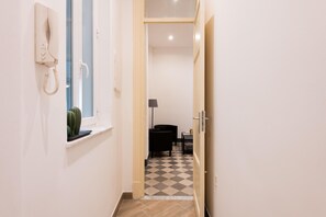 Ein schmaler Flur mit Telefon an der Wand, Fenster mit einem Kaktus, schwarzem Stuhl, Lampe und kariertem Boden.