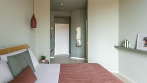 Ein Schlafzimmer mit einem Bett, einer Zimmerpflanze, einer roten Pendelleuchte und einem zur Toilette führenden Flur.