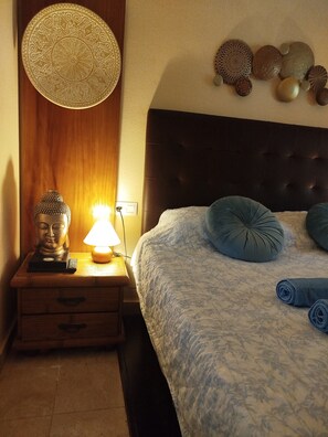 Una cama bien tendida con un edredón estampado, una mesita de noche de madera con una estatua de Buda y una lámpara, y una pared decorada con placas.