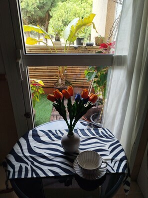Una ventana con vistas a un jardín, una mesa con mantel a rayas, un jarrón con flores y un cuenco.
