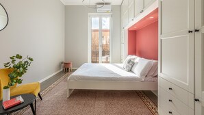 Ein Schlafzimmer mit einem Bett, einem Sessel, einem kleinen Tisch, einer Topfpflanze und Blick ins Freie.