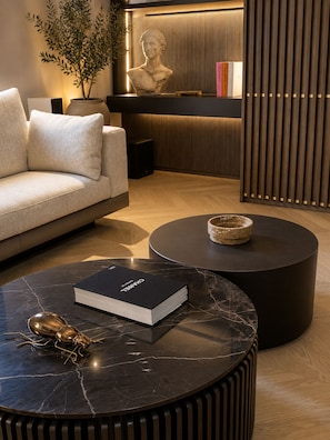 A modern living room with a sofa, a marble coffee table with a book and a decorative insect, a round side table, a potted plant, and a bust sculpture against a wooden backdrop. - ASTRA PENTHOUSE (Bologna)