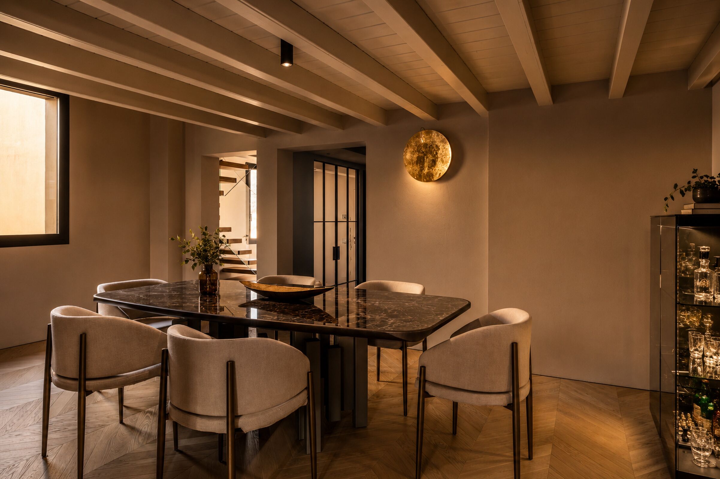 A dining area with a round table and six chairs, a wall-mounted light fixture, and a glass cabinet with bottles.