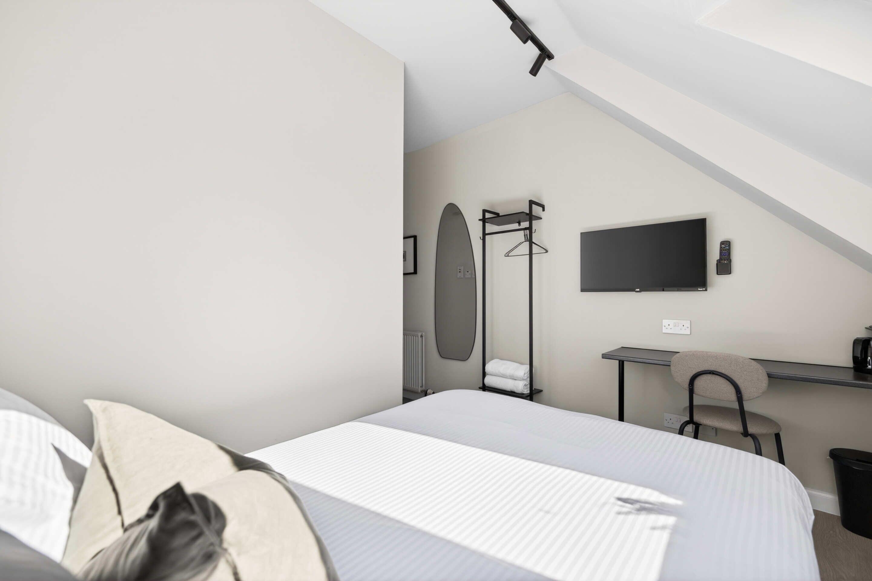 A modern hotel room with a bed, a desk, a chair, a TV, and a coat rack.