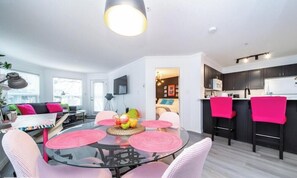 A modern living room with a glass dining table, pink chairs, and a kitchen with dark cabinets and a white countertop. - Luxury Duo Suites - King Bed - AC - Patio - UGPark (Edmonton)