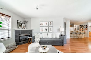 A modern living room with a fireplace, grey sofa, and a dining area with chairs. - Luxury Duo Suites - King Bed - AC - Patio - UGPark (Edmonton)