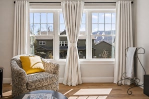 A room with a patterned armchair, a wooden floor, a window with curtains, and a towel rack. - Summit Ridge - New 5BR Home in Lincoln - Near Trails, River & Loon (Lincoln)