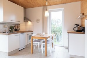 Eine moderne Küche mit weißen Schränken, einem Holztisch und Blick auf Grün hinter dem Fenster.