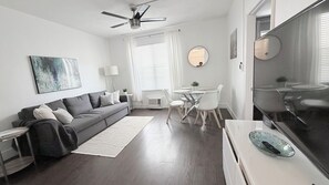 A modern living room with a grey sofa, a dining table with chairs, a TV, and a ceiling fan. - 1 BR on Collins Ave. steps away from the BEACH (Miami)