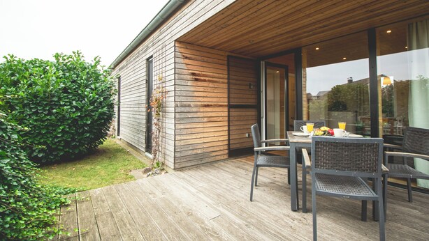 Een houten terras met een tafel en stoelen, een glazen deur naar een kamer, en een groene haag.