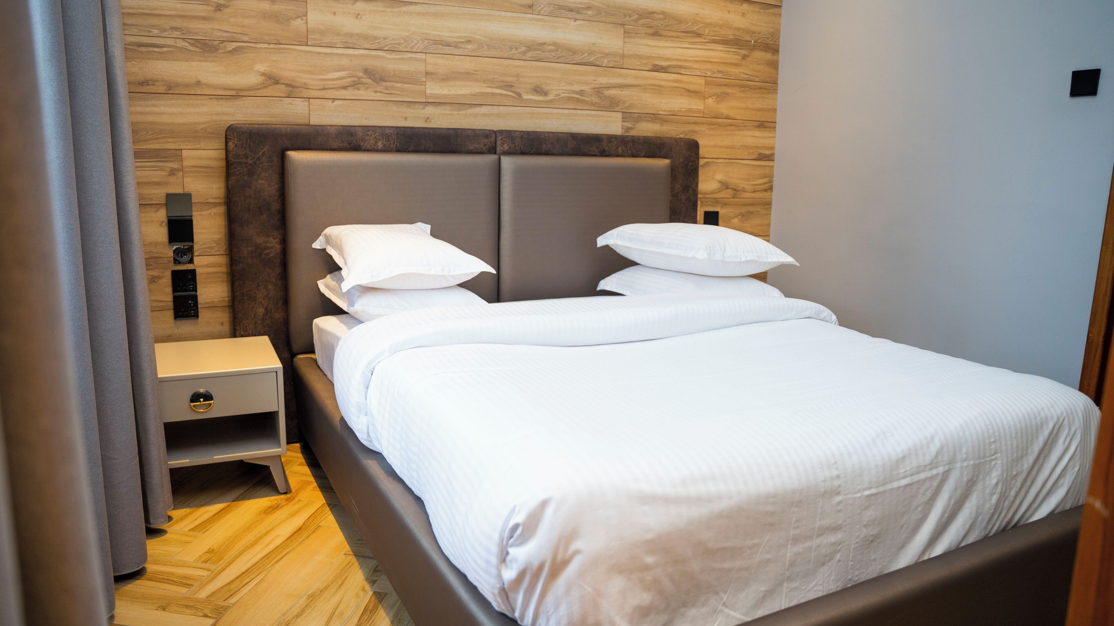 A neatly made bed with white linens, a wooden headboard, a bedside table with a drawer, and a wall-mounted power outlet.