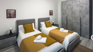 Et moderne hotelværelse med to senge, et garderobeskab, et natbord med lampe og en stol i badeværelset.