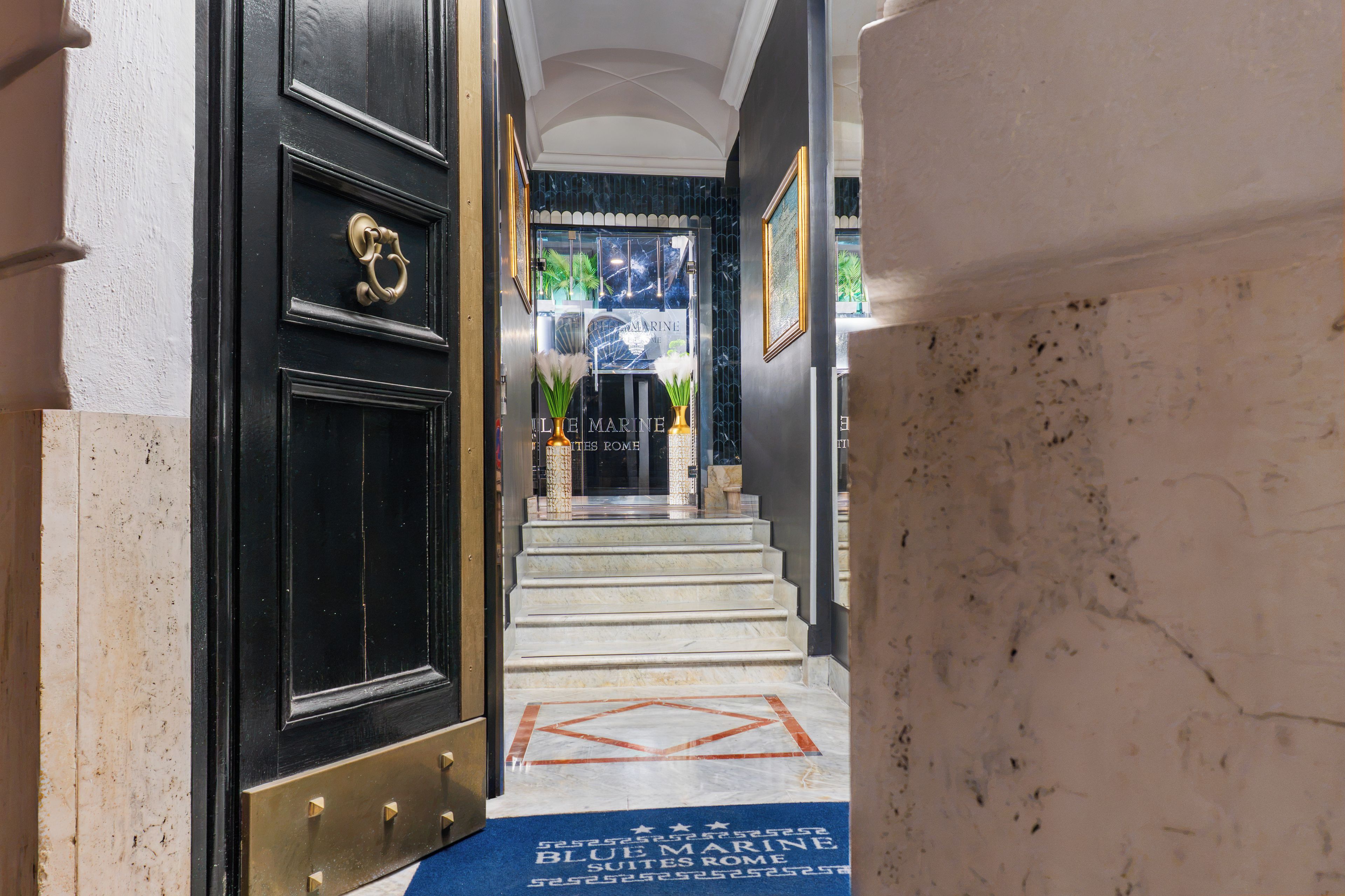 Una escalera de mármol conduce a una puerta negra con un llamador de latón, una alfombra azul con texto y un cuadro en la pared.