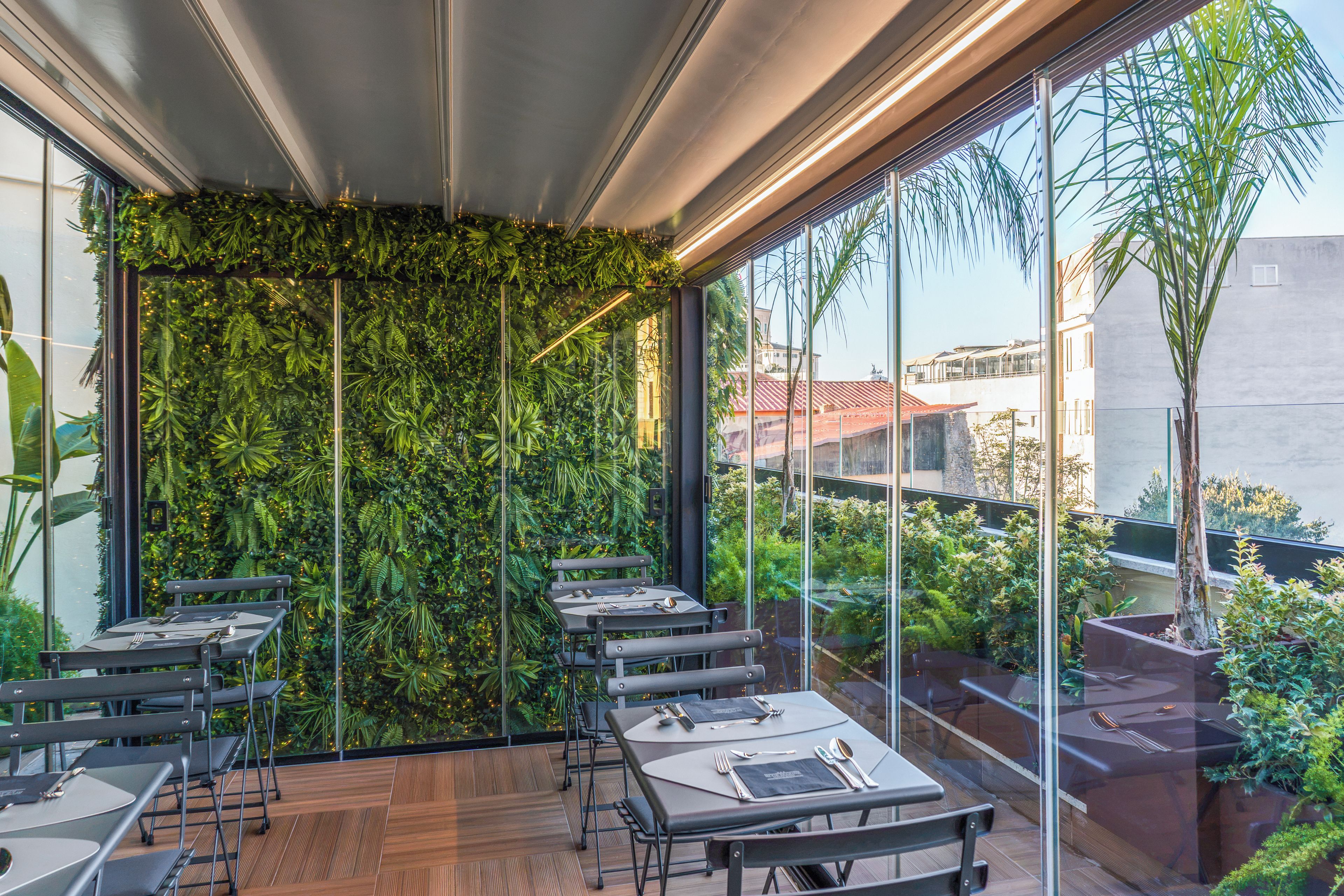 Un restaurante con mesas dispuestas para comer, una pared verde con plantas y vistas a edificios a través de amplios ventanales.