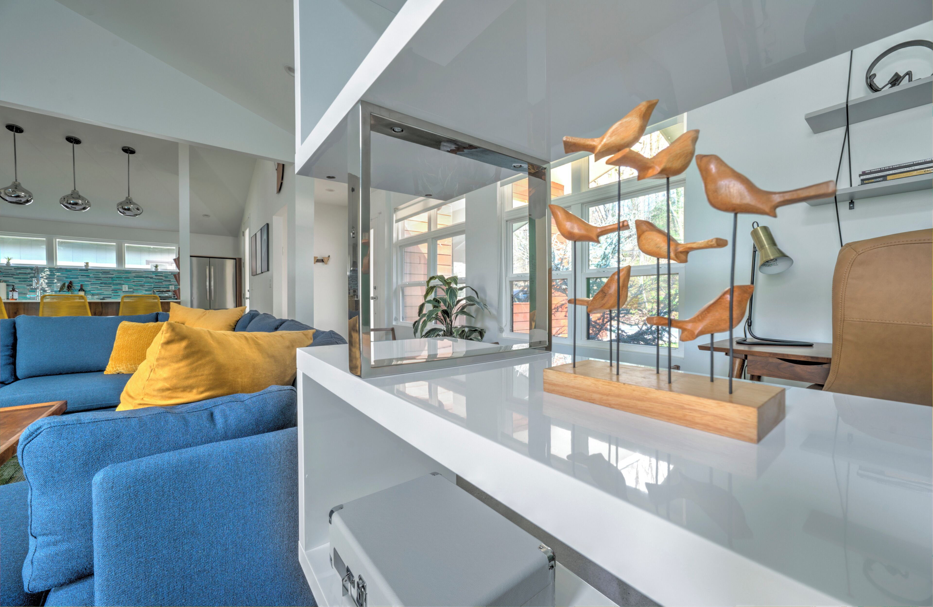 Un salon moderne avec un canapé bleu, des coussins jaunes, une table basse en verre et des sculptures d’oiseaux en bois.