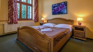 A wooden bed with white linens, two bedside tables with lamps, a window with curtains, and a floral painting on the wall.