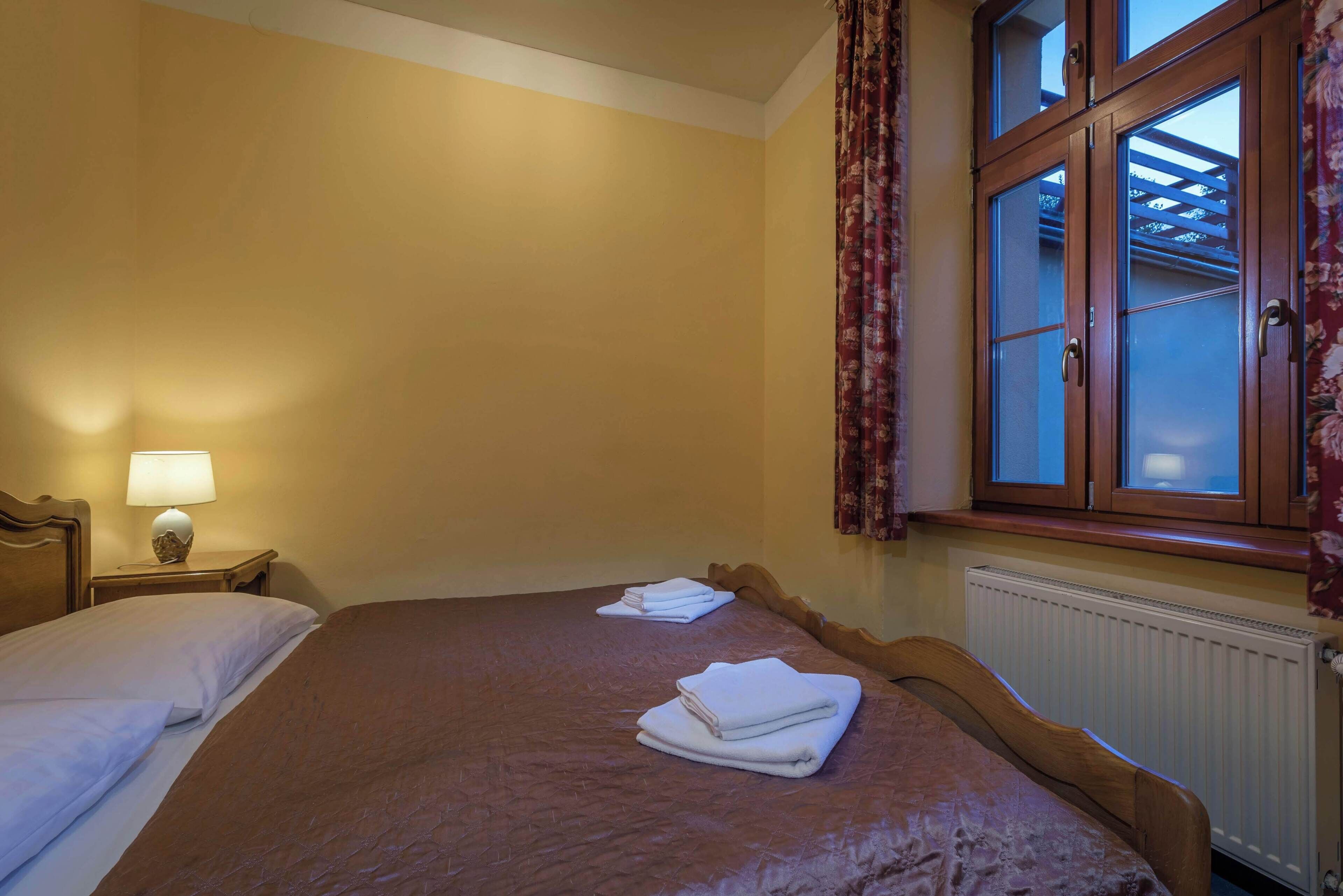 A hotel room with two beds, a nightstand with a lamp, a window with curtains, and a radiator.